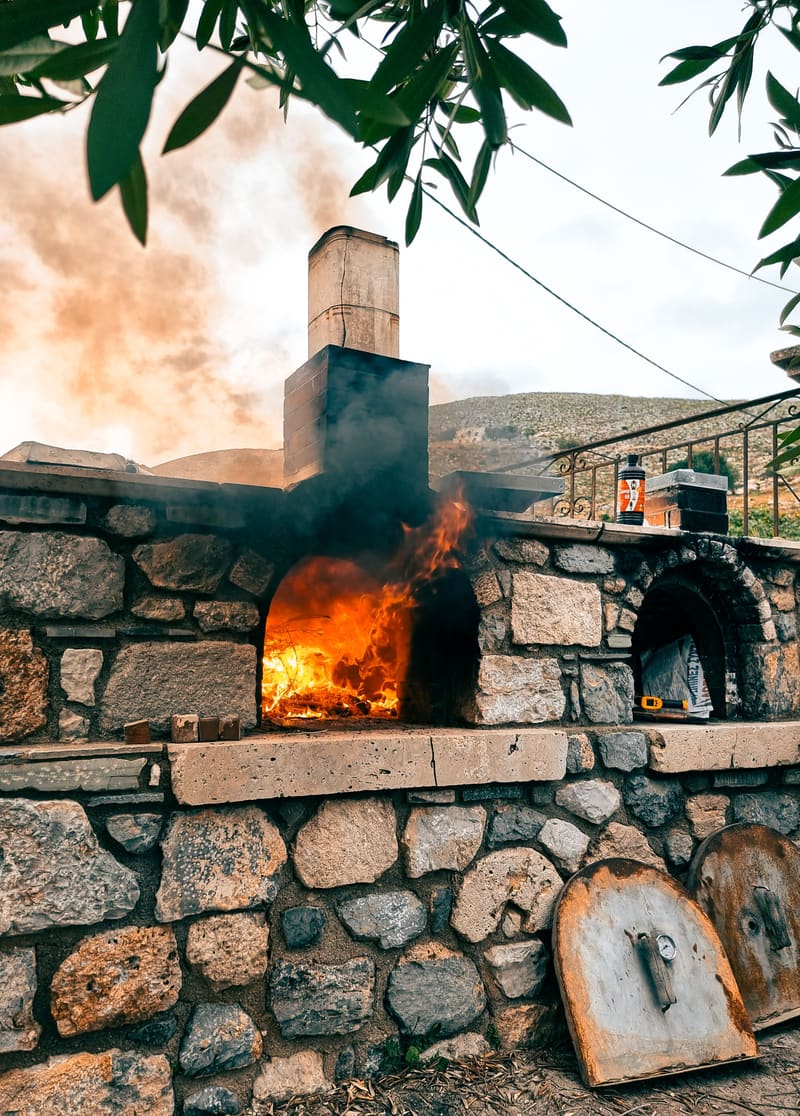 A wood-fired stone oven burning beside a stone wall, smoke rising.