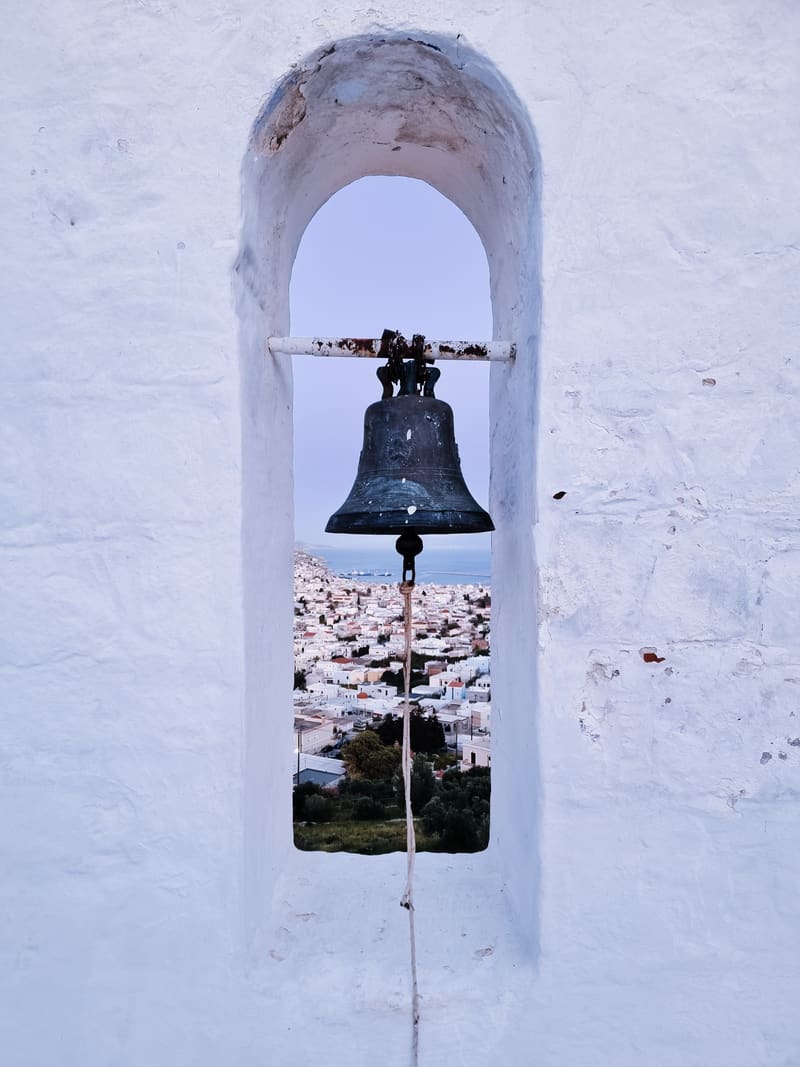 A church bell hanging in an arched niche in a whitewashed wall, with a white village visible through the opening.