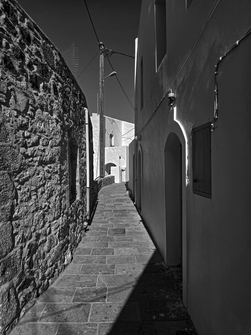 Black and white: a narrow stone-walled alley with a strip of sunlight on the paving.