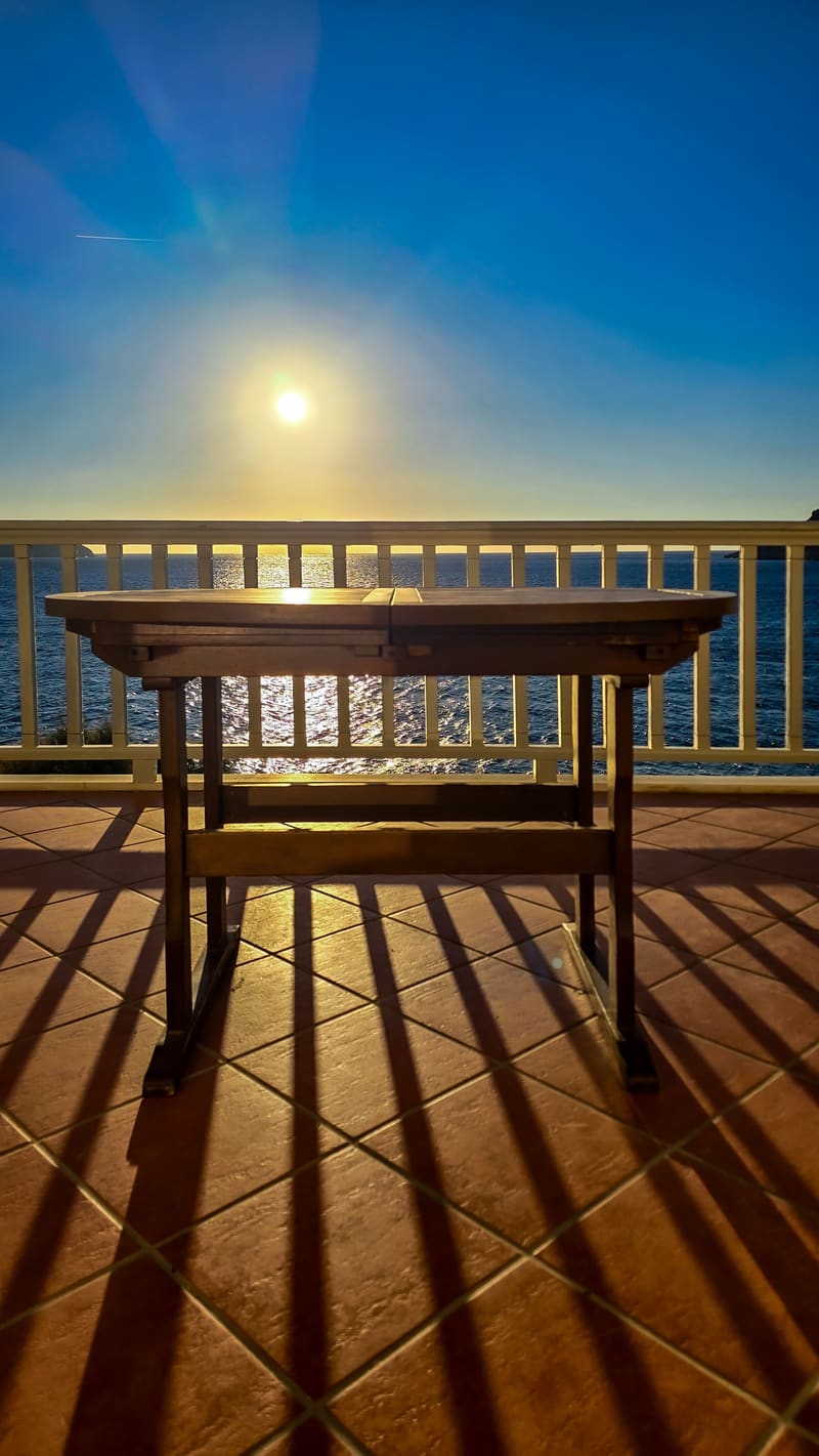 A wooden table on a balcony facing the sun over the sea.