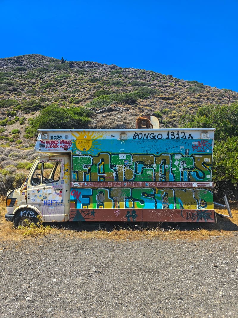 An old van covered in graffiti, parked on gravel below a scrubby hillside.