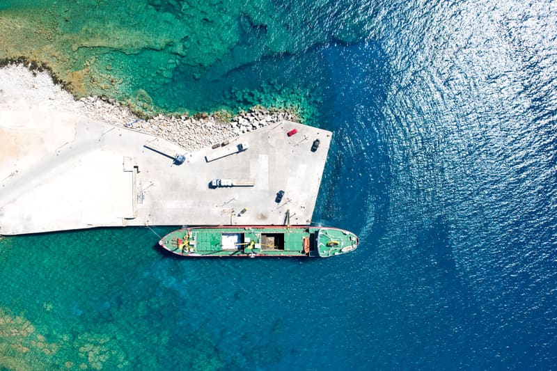 Aerial view of a cargo ship moored at a concrete pier, turquoise shallows meeting deep blue sea.