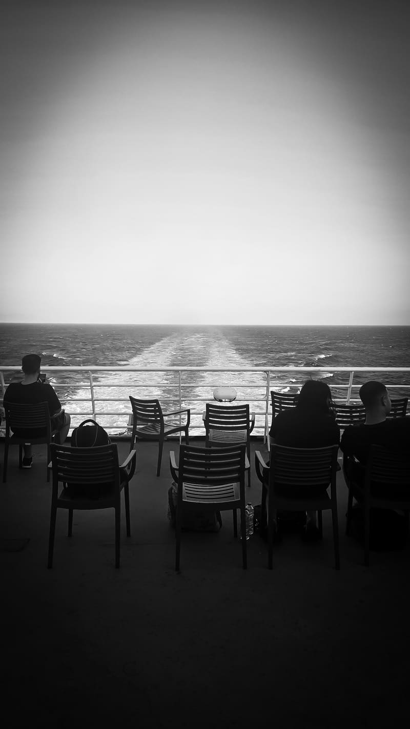 Black and white: passengers in silhouette on a ferry's rear deck, facing the wake and the bright sea.