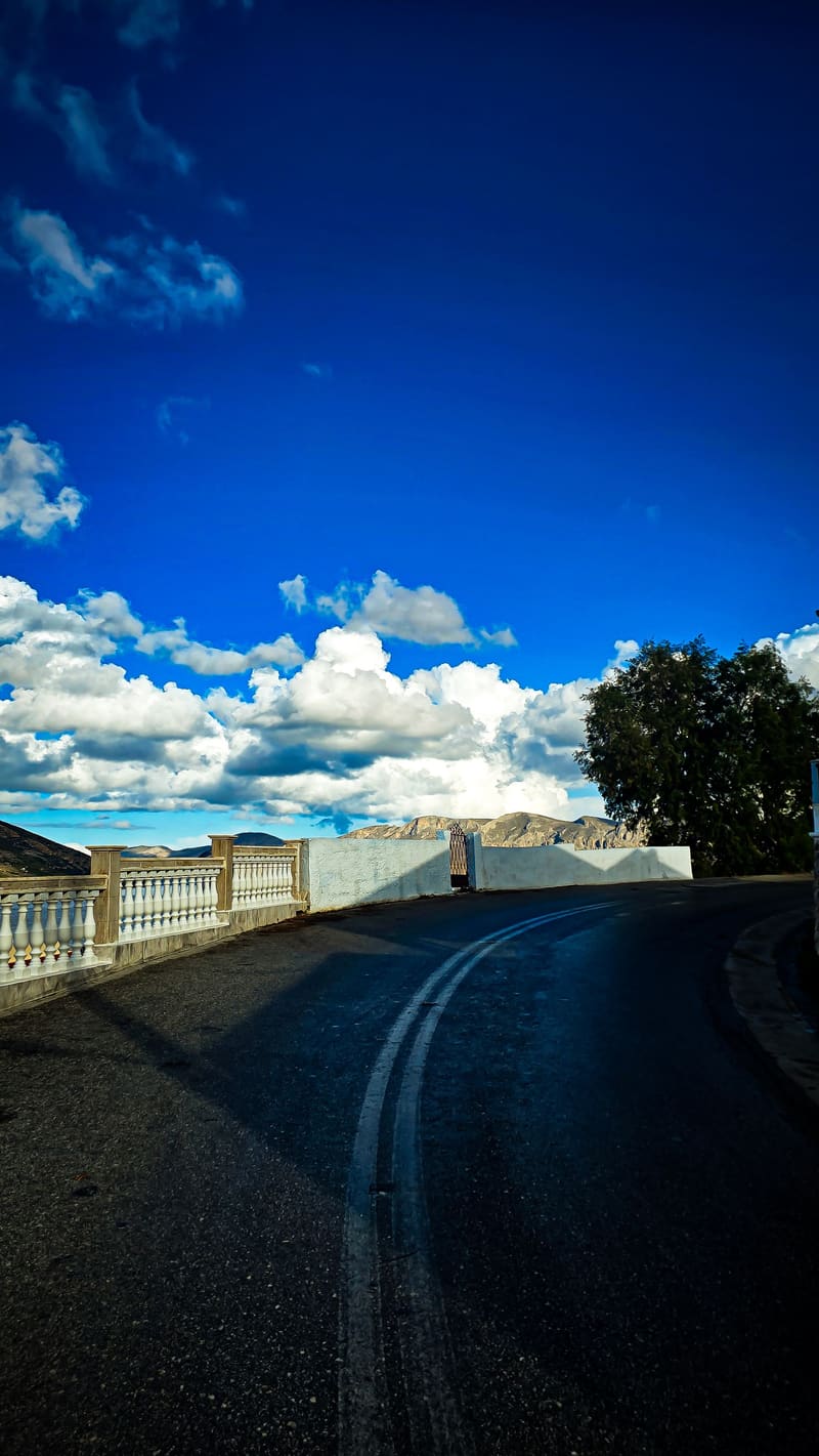 A winding road beside a white balustrade under a blue sky and clouds.