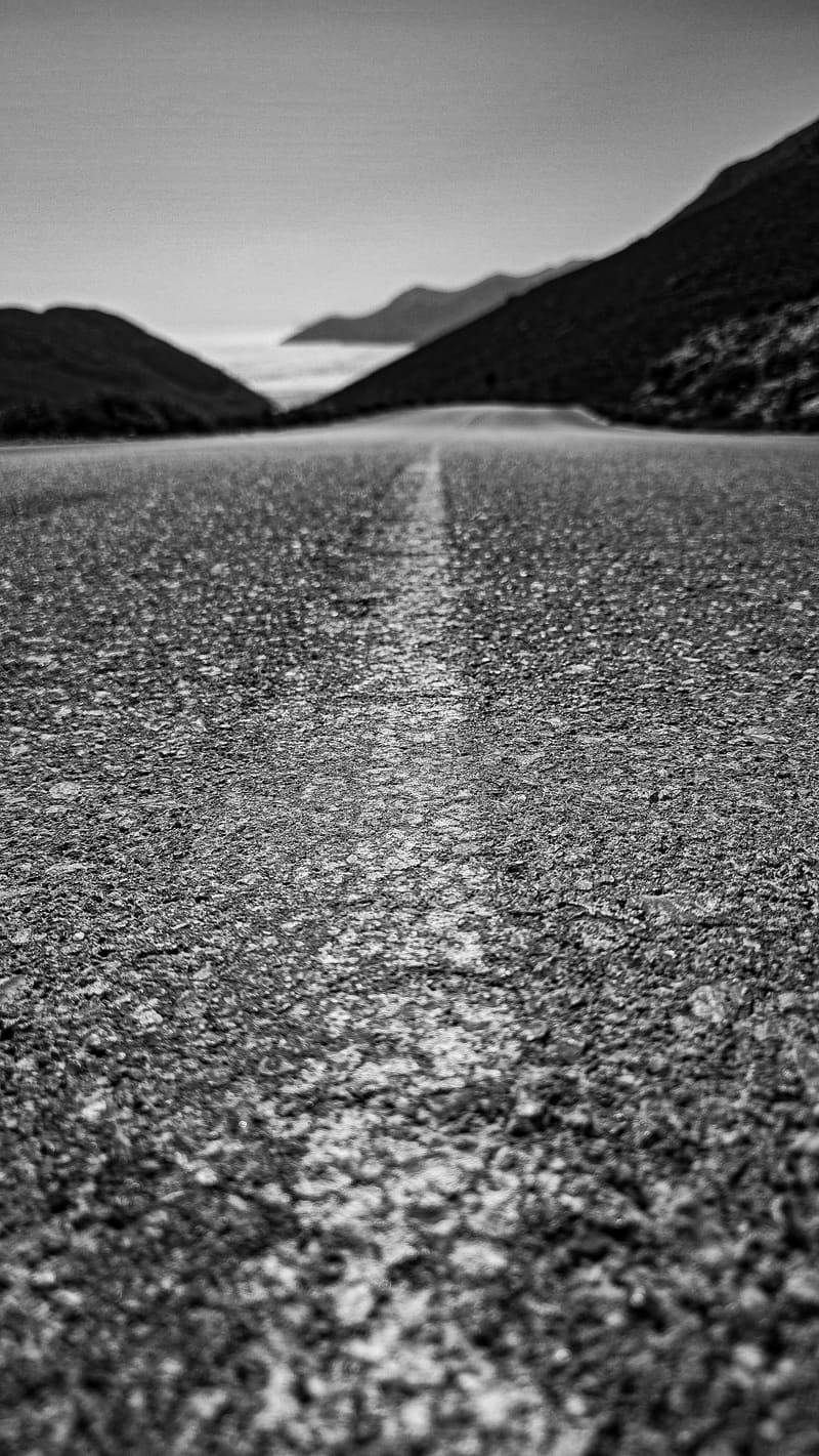 Black and white: a road seen from ground level, its centre line running towards distant mountains.