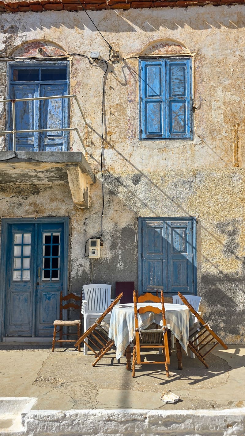 A weathered house front with blue shutters and doors, café tables and chairs outside.