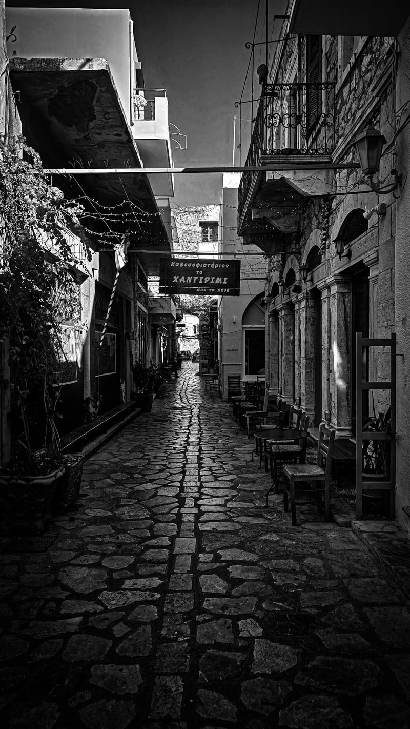 Black and white: a cobbled street between old buildings with balconies and hanging signs.