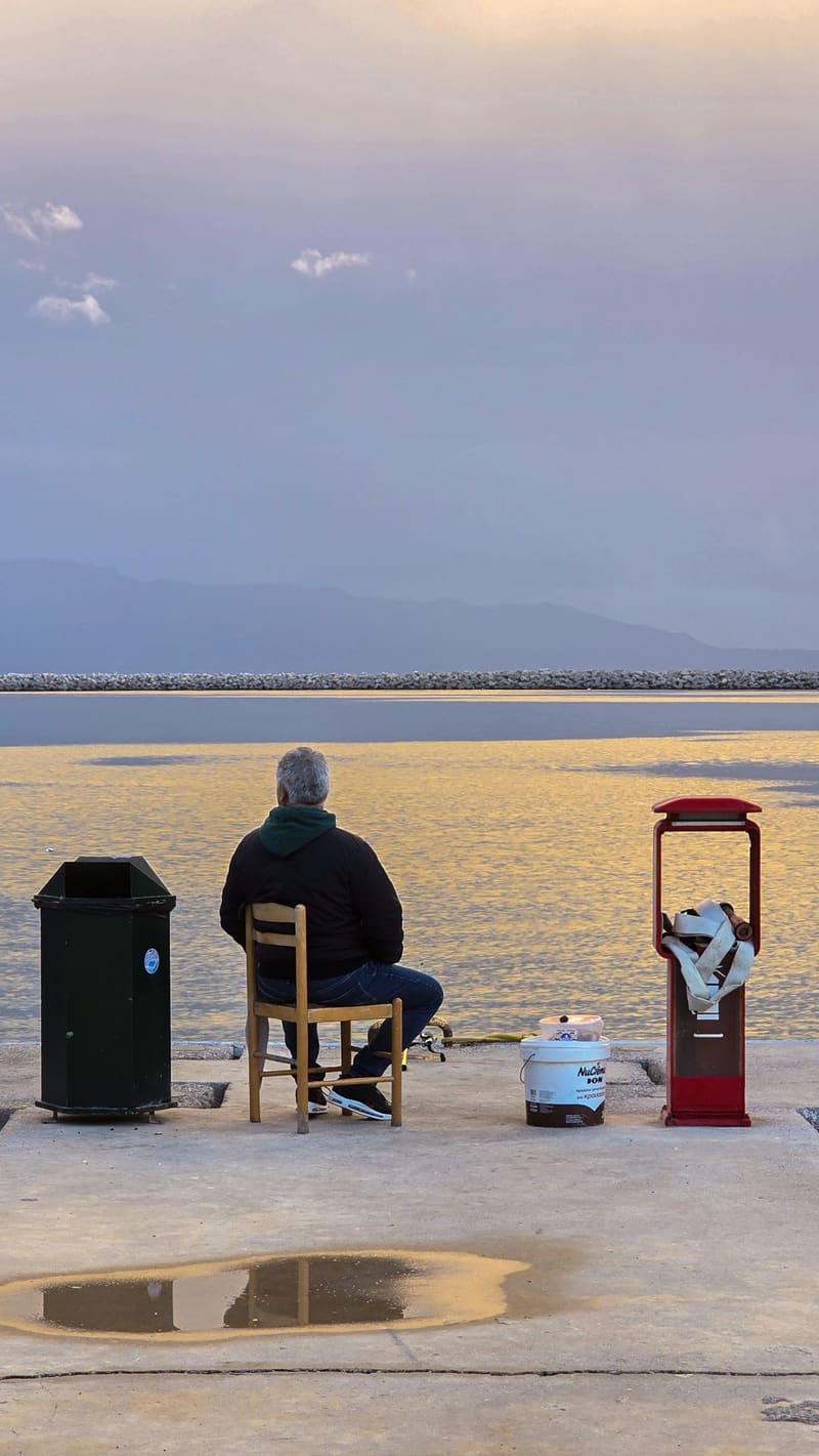 A man fishing from a chair on a quayside at dusk, seen from behind, a puddle reflecting the sky.