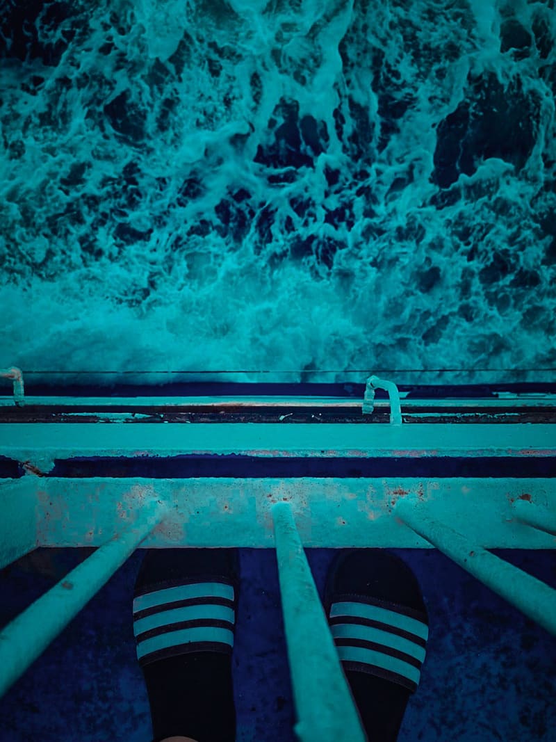 Looking down from a ship's railing at churning turquoise water, feet in striped socks at the bottom of the frame.