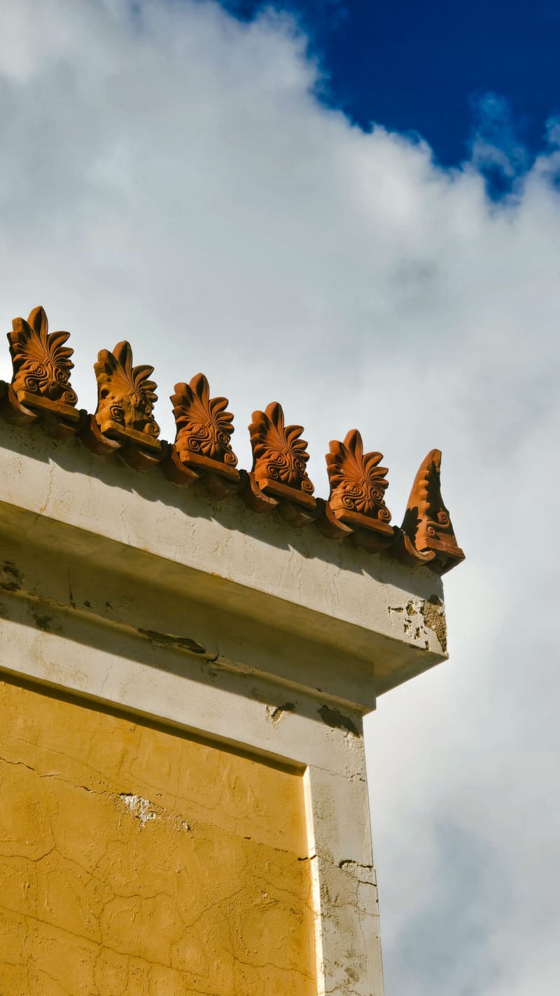 The corner of a yellow building topped with terracotta ornaments, against blue sky and cloud.