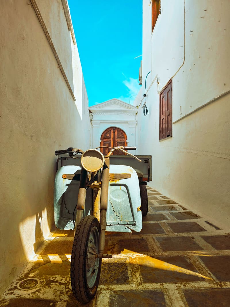 An old motorbike with a sidecar parked in a narrow whitewashed lane, under a strip of blue sky.