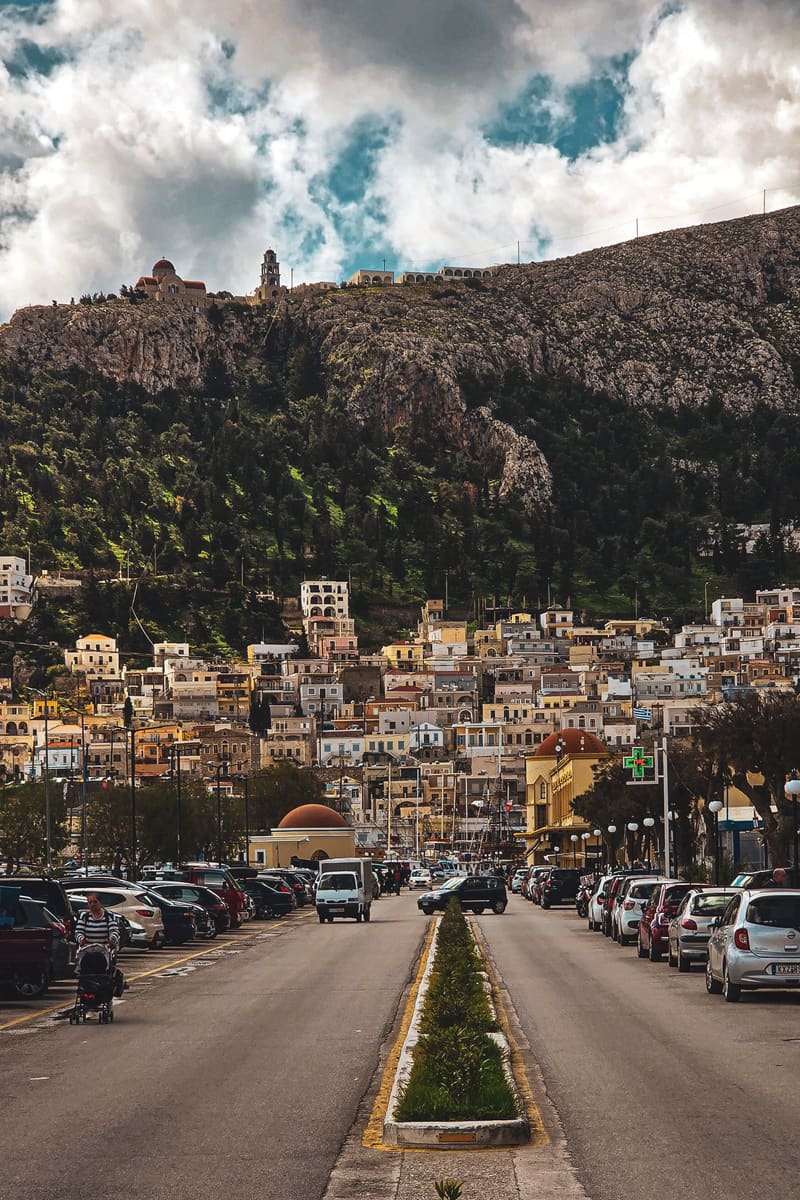 A long avenue lined with parked cars, leading to a hillside town.