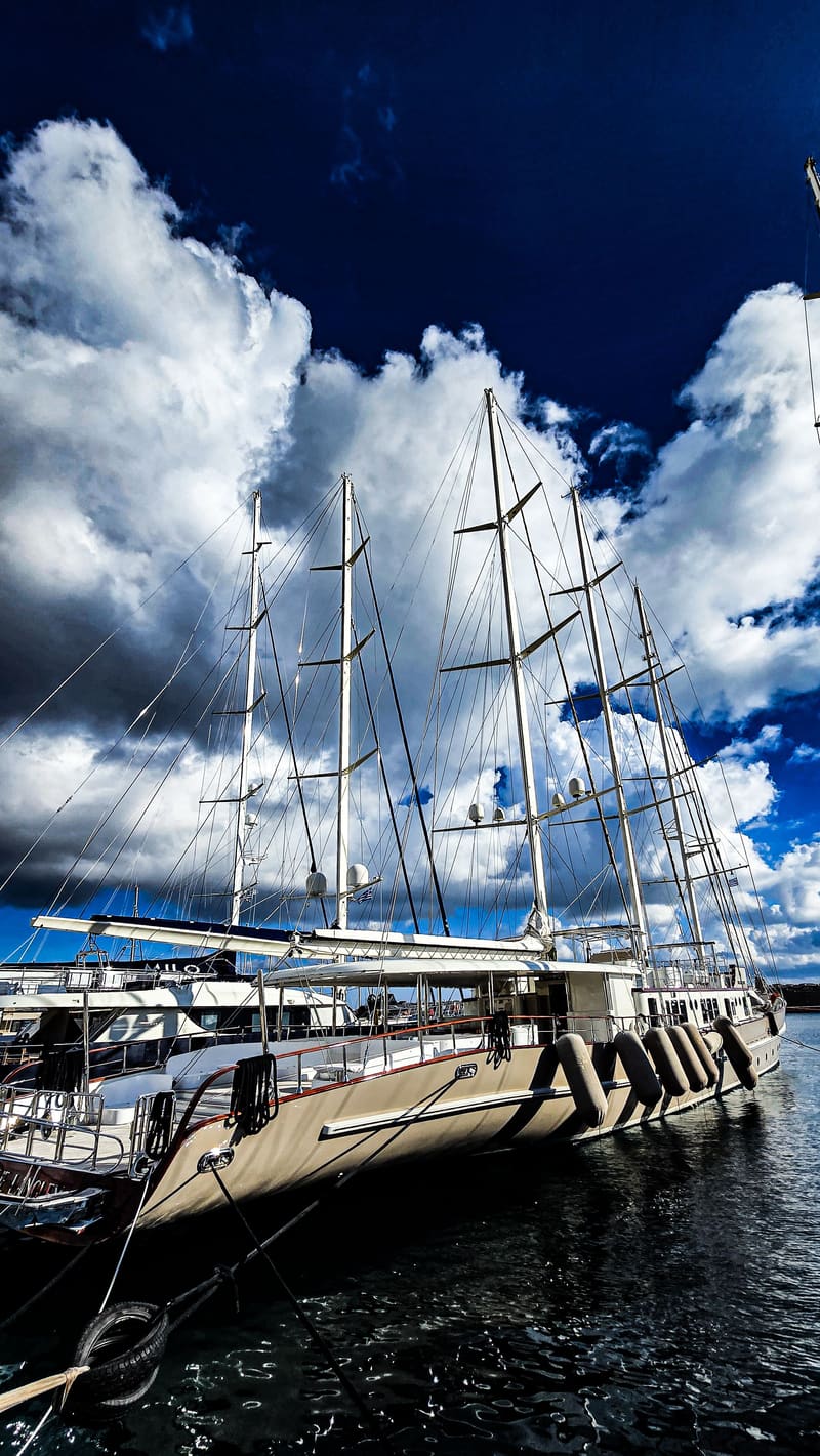 A tall sailing ship moored in a harbour, its masts against dramatic white clouds.