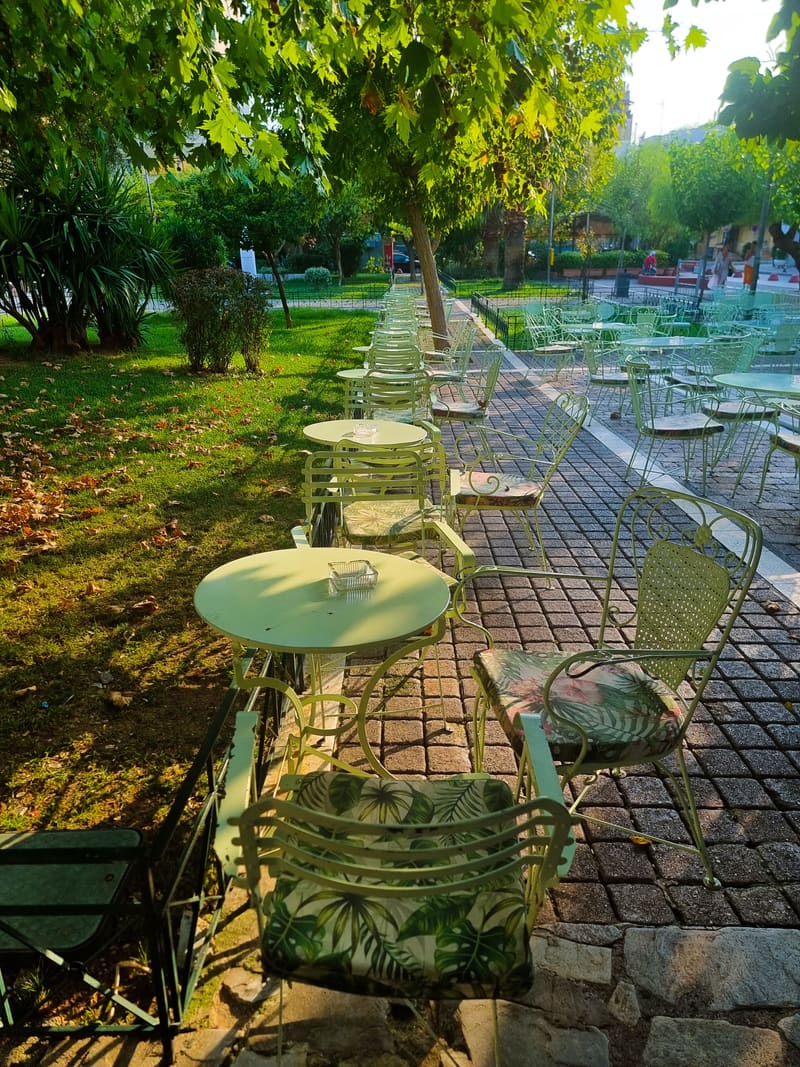 Empty café tables and chairs under trees along a paved path.