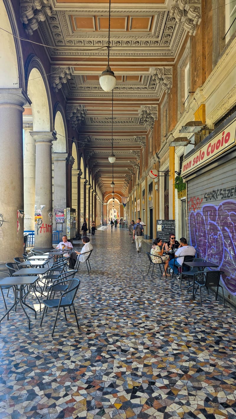 A long arcade with a painted ceiling and patterned stone floor, café tables along one side.