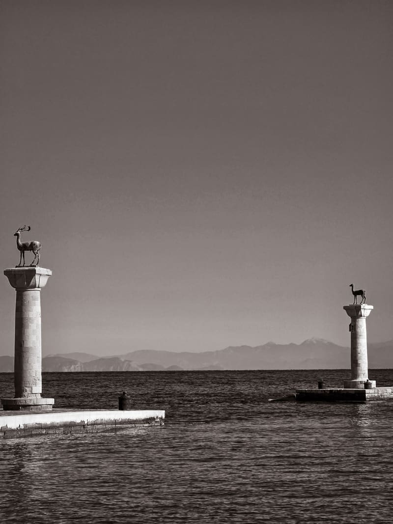 Black and white: two columns topped with deer statues at a harbour entrance, mountains beyond.