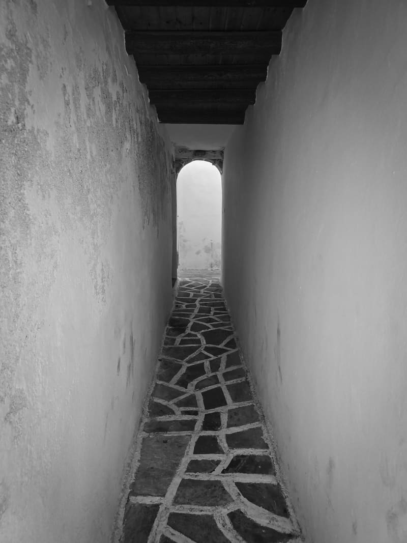 Black and white: a narrow whitewashed passage with a stone-paved floor, leading to a bright arched opening.