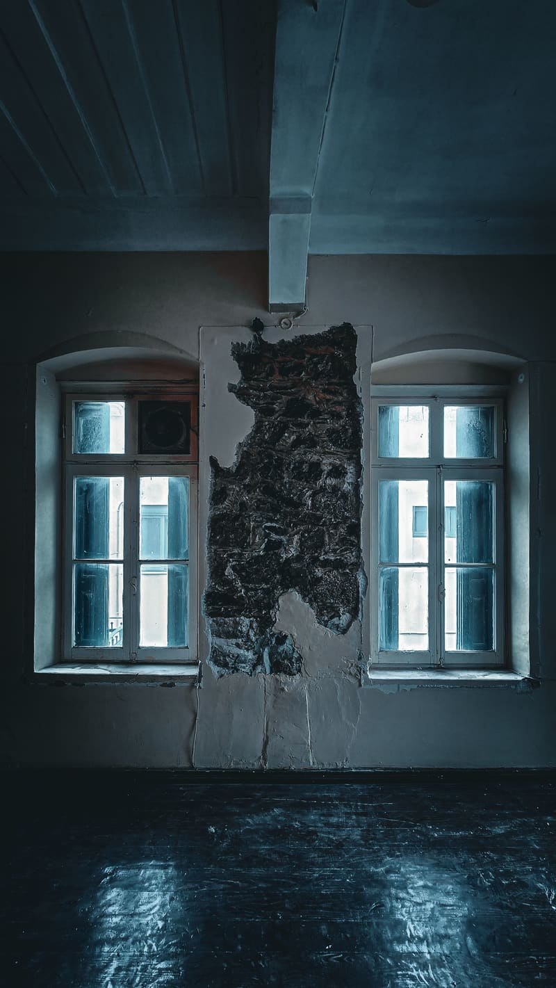 An empty room with two windows on either side of a patch of bare, peeling wall.