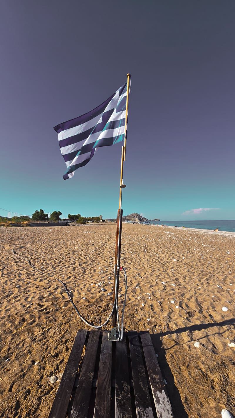A Greek flag on a pole above a sandy beach.