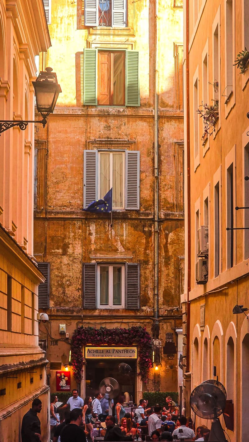 A narrow street of tall ochre buildings with a busy restaurant below.