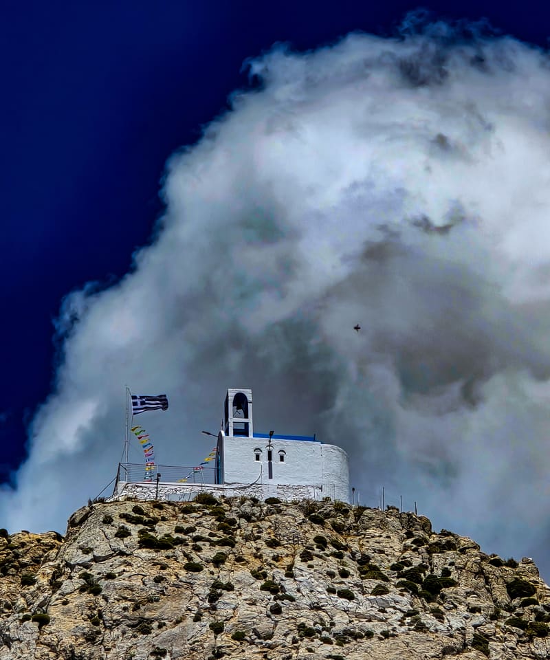 A small white chapel and Greek flag on a rocky peak, under a towering white cloud.