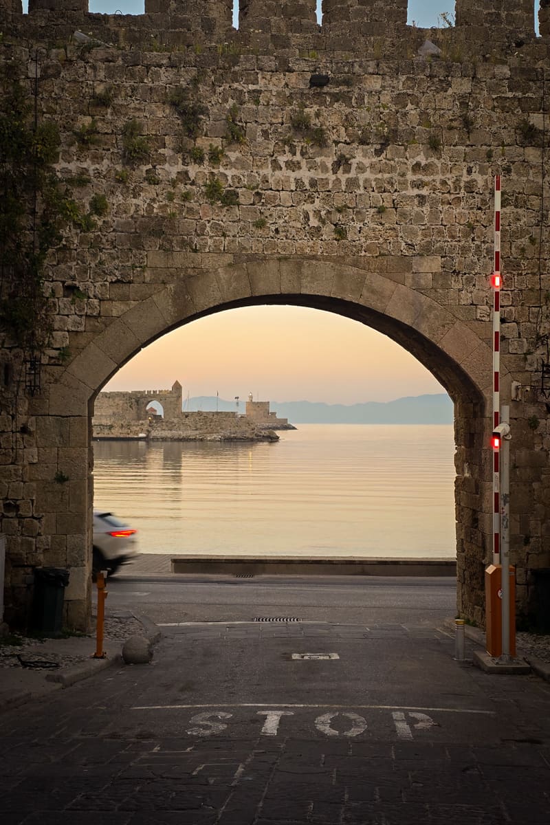 A stone gateway in a medieval wall, framing the sea and a harbour at dusk; STOP painted on the road.