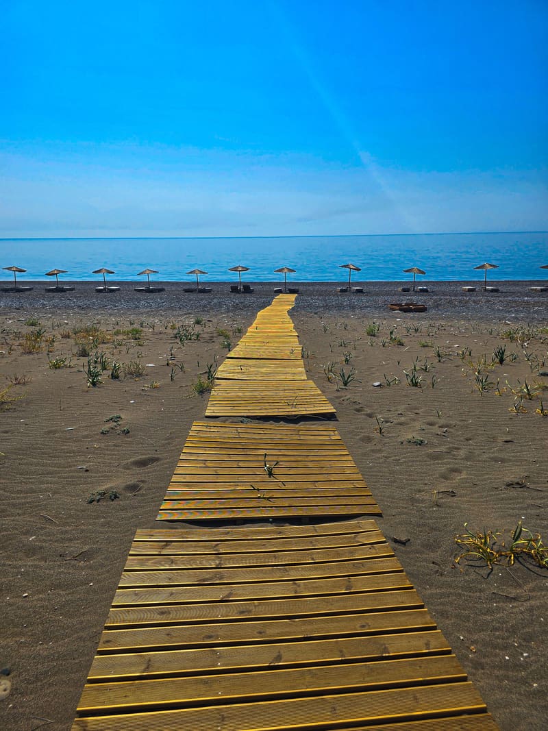 A yellow wooden boardwalk crossing dark sand towards the sea and a row of umbrellas.
