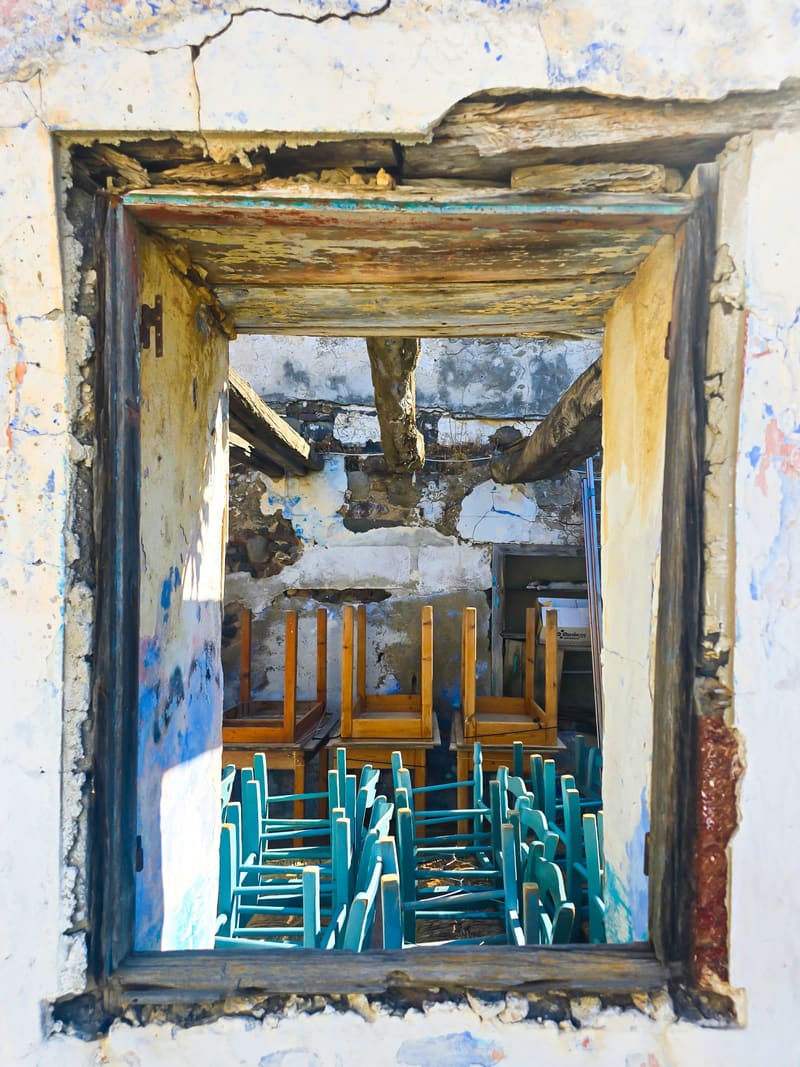 A weathered window frame in a crumbling wall, with stacked turquoise and wooden chairs inside.