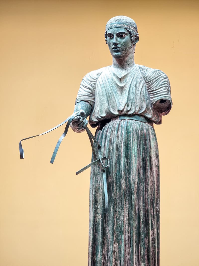 A bronze statue of a robed charioteer holding reins, against a warm ochre wall.