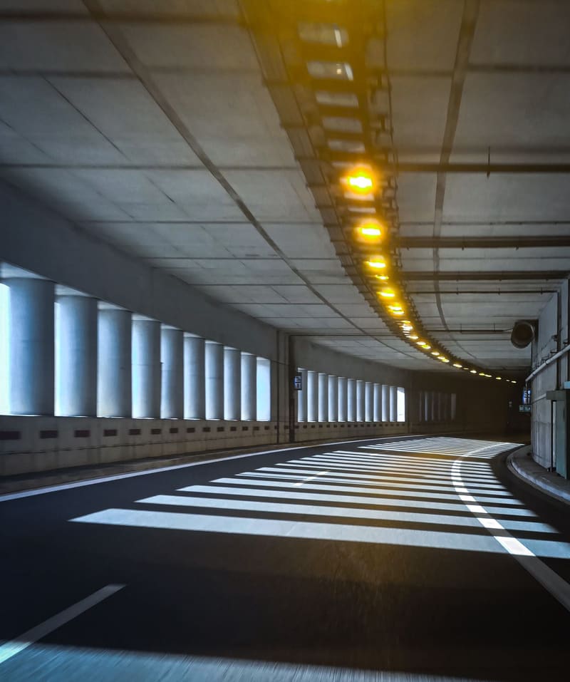 A covered road under a concrete ceiling, its amber lights curving away, sunlight striping the floor.