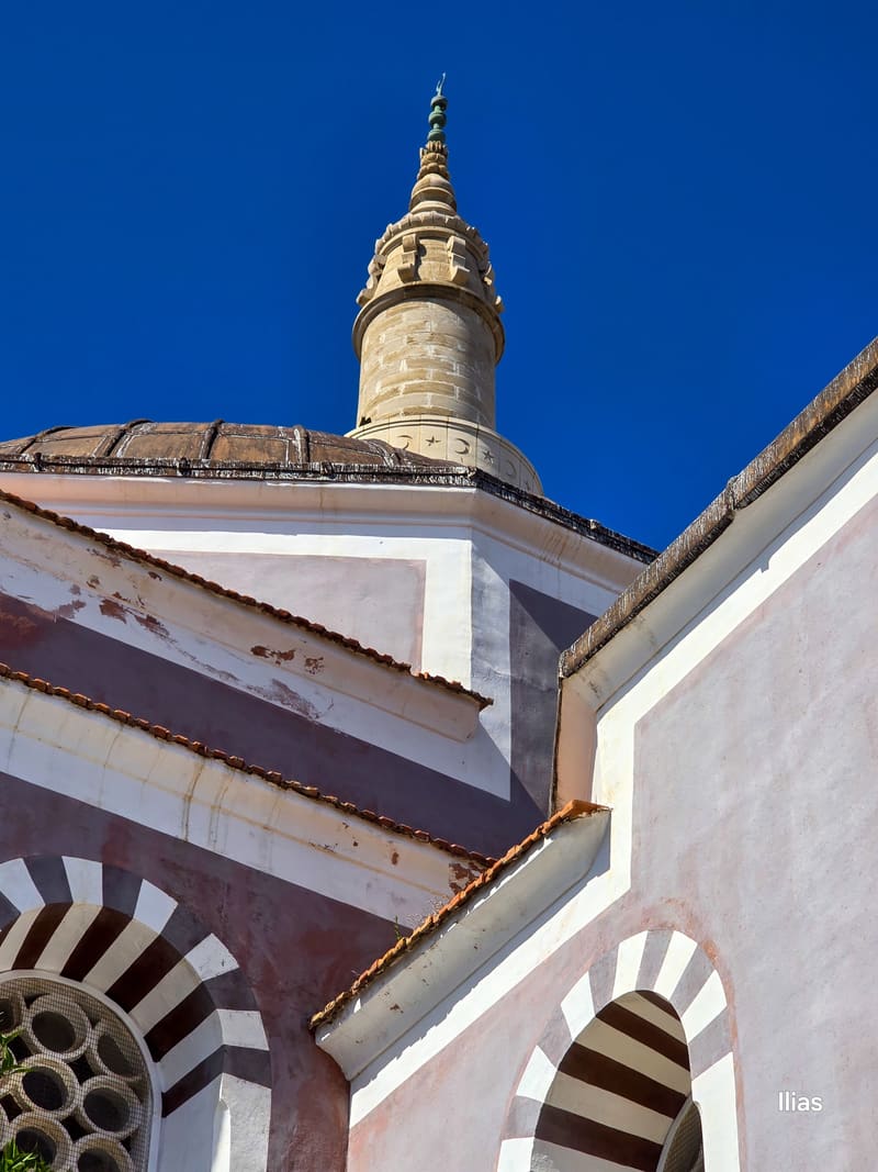 A dome and a slender tower above striped arches, against a blue sky.