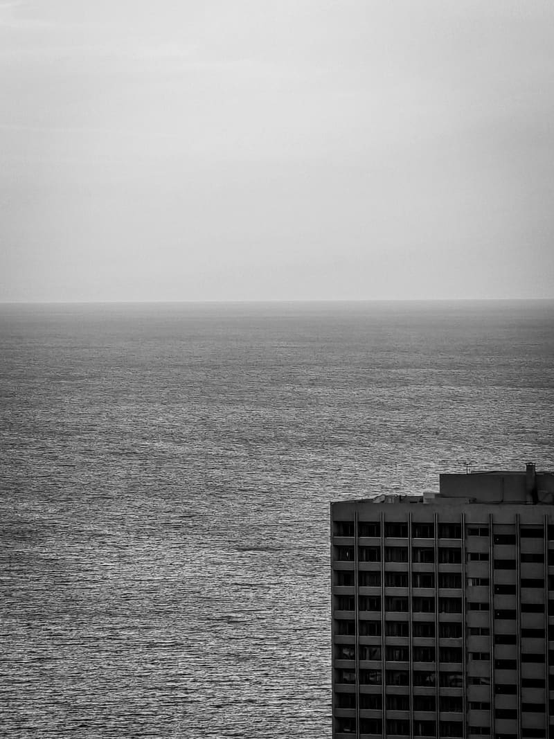 Black and white: a calm sea beside the edge of a tall building.