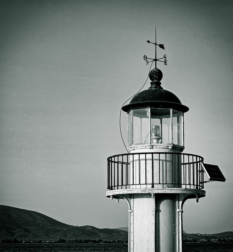 Black and white: the lantern and railed gallery at the top of a lighthouse, with hills behind.