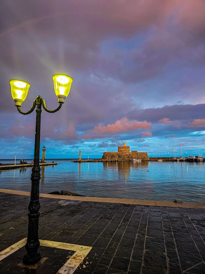 A double street lamp lit on a harbour front at dusk, a fortress across the water.