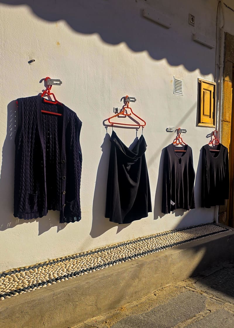 Black garments on red hangers along a sunlit white wall, under the shadow of an awning.