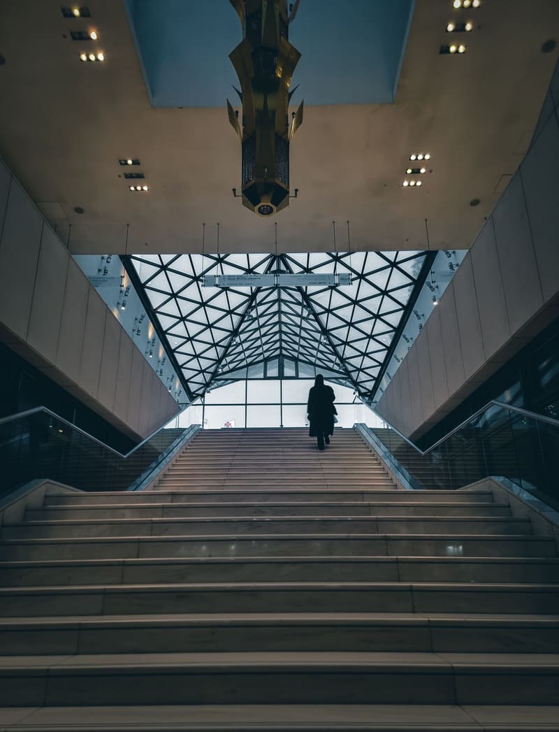 A wide staircase climbing towards a glass roof of triangular panes, one figure at the top.