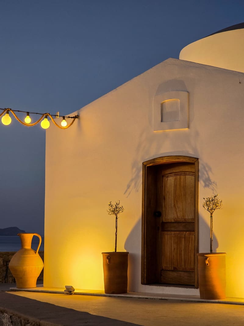 A small white chapel with a wooden door, lit warm yellow at dusk, a string of bulbs above.