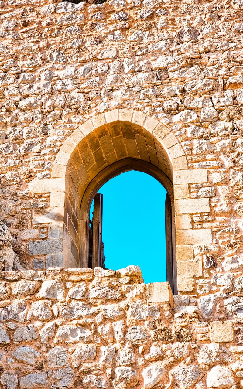 An arched window in a stone fortress wall, framing a patch of bright blue sky.