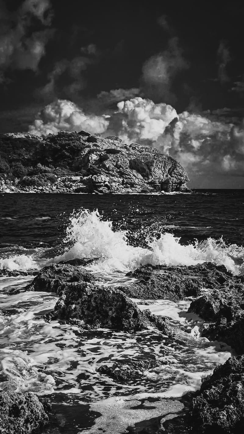 Black and white: waves breaking over dark rocks beneath a headland and heavy clouds.