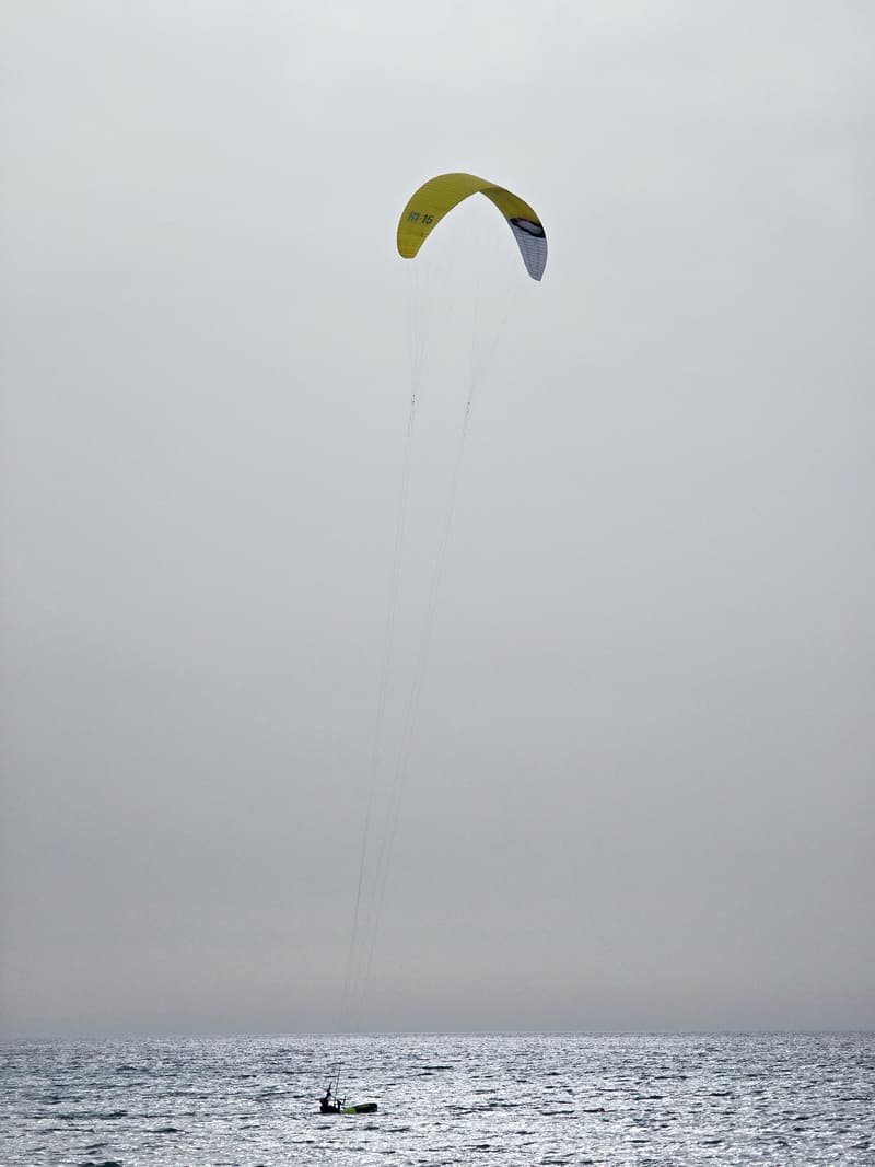 A lone kitesurfer on a grey sea, the kite small against a pale, empty sky.