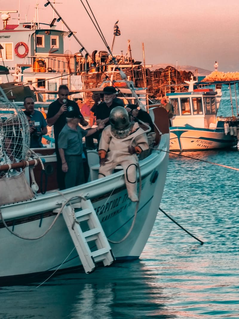 Fishermen on a boat in a harbour, one in an old-style diving suit, with fishing boats behind.