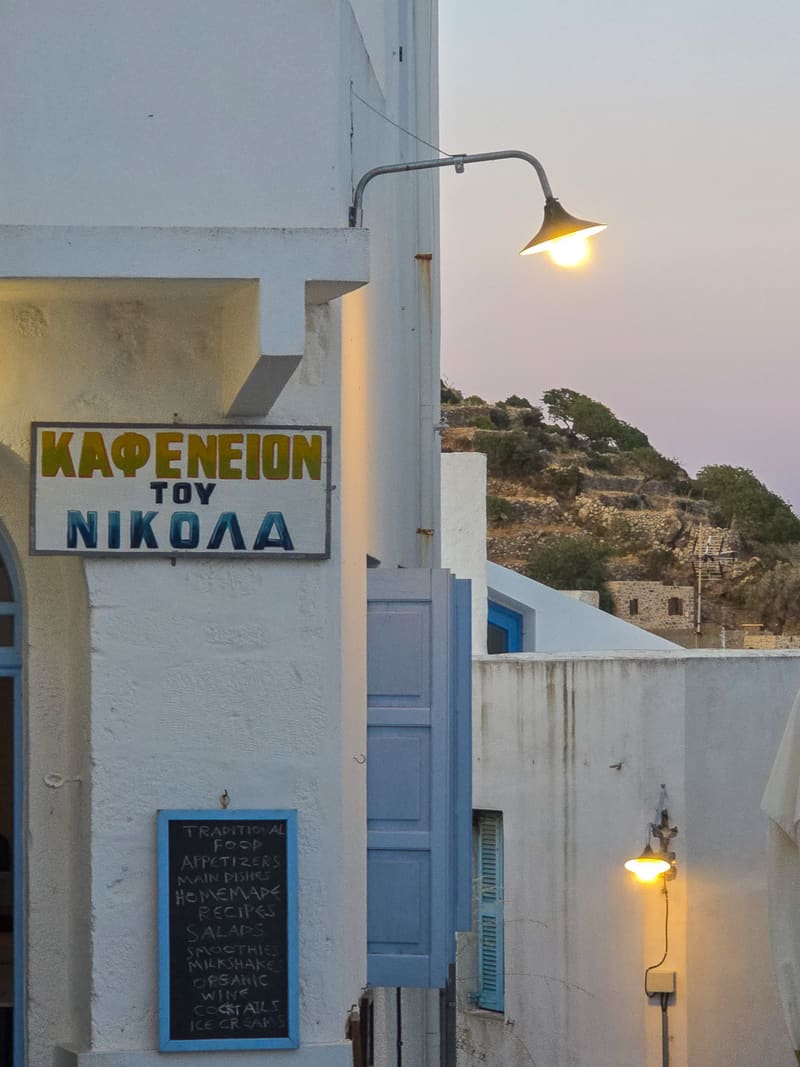 A white café wall with a sign reading "Καφενείον του Νικόλα" and a lit lamp.