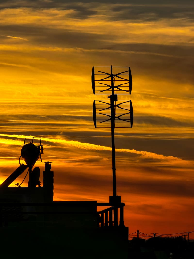 An antenna and rooftops in silhouette against a blazing orange sunset.