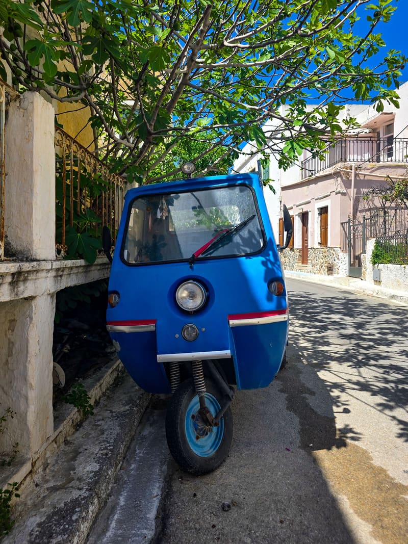 A blue three-wheeled vehicle parked in a sunny village lane under a tree.
