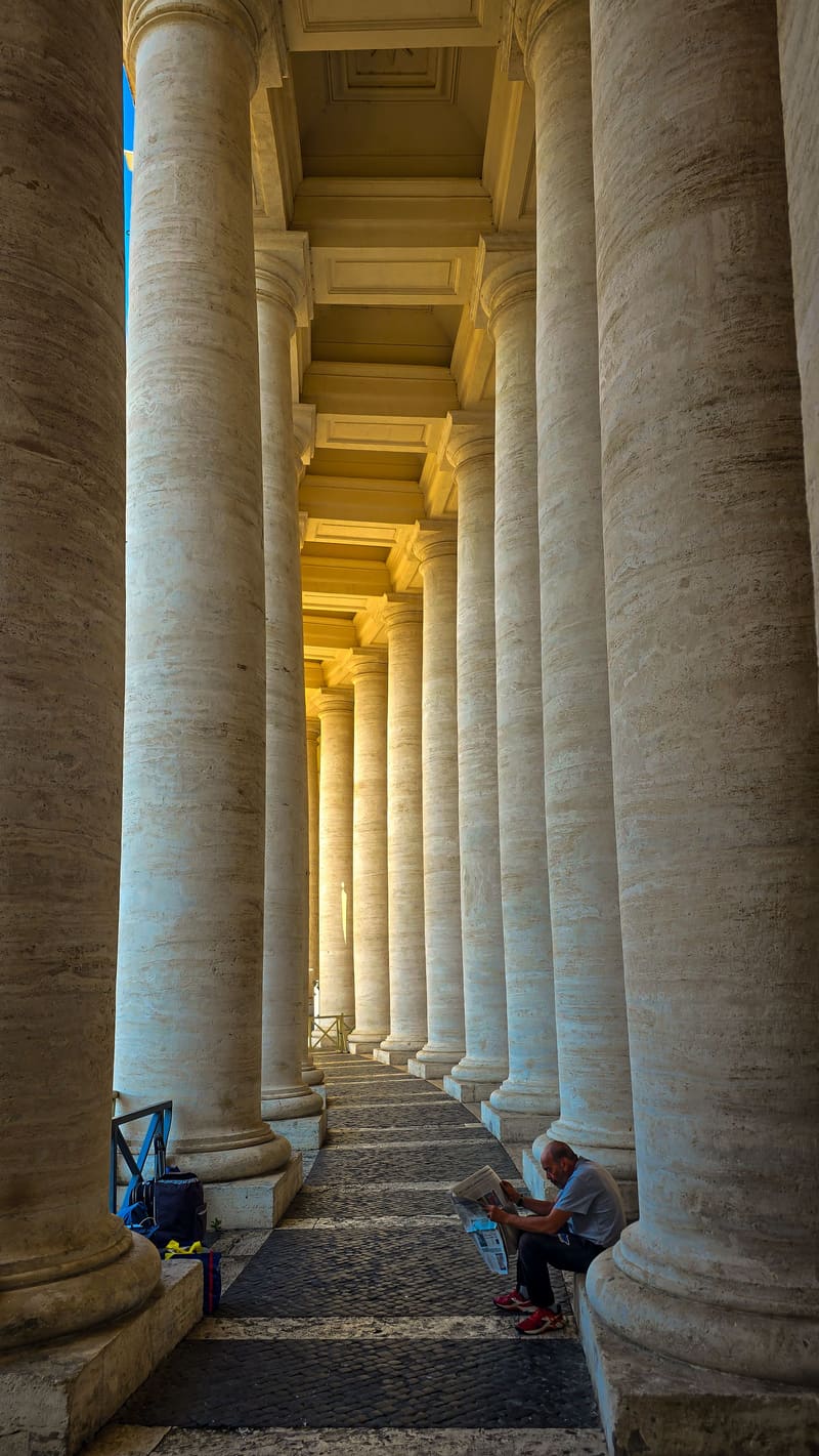 A colonnade of tall stone columns receding into the distance, two people sitting at the base.