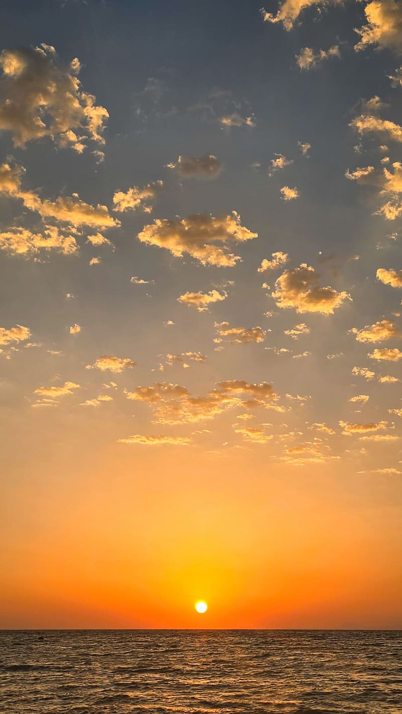 The sun setting into the sea under scattered golden clouds.