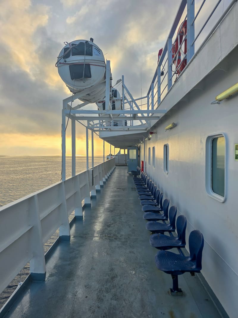 The empty side deck of a ferry, a row of blue seats leading towards the sea and a lifeboat.