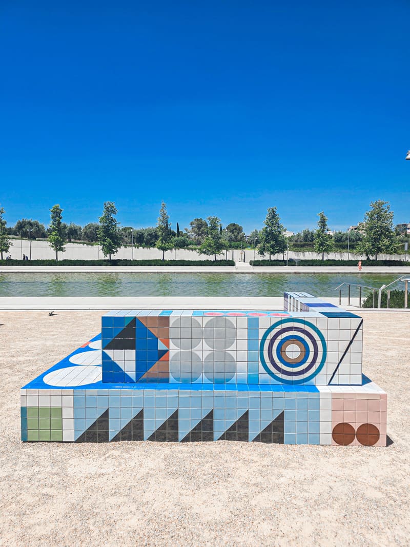 A bench of geometric tiles in blue, white and grey on a sandy square, trees behind.