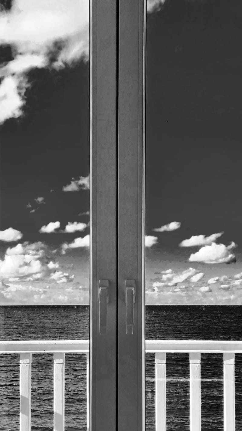 Black and white: clouds and sea seen through a glass door, divided by its frame and a balcony railing.